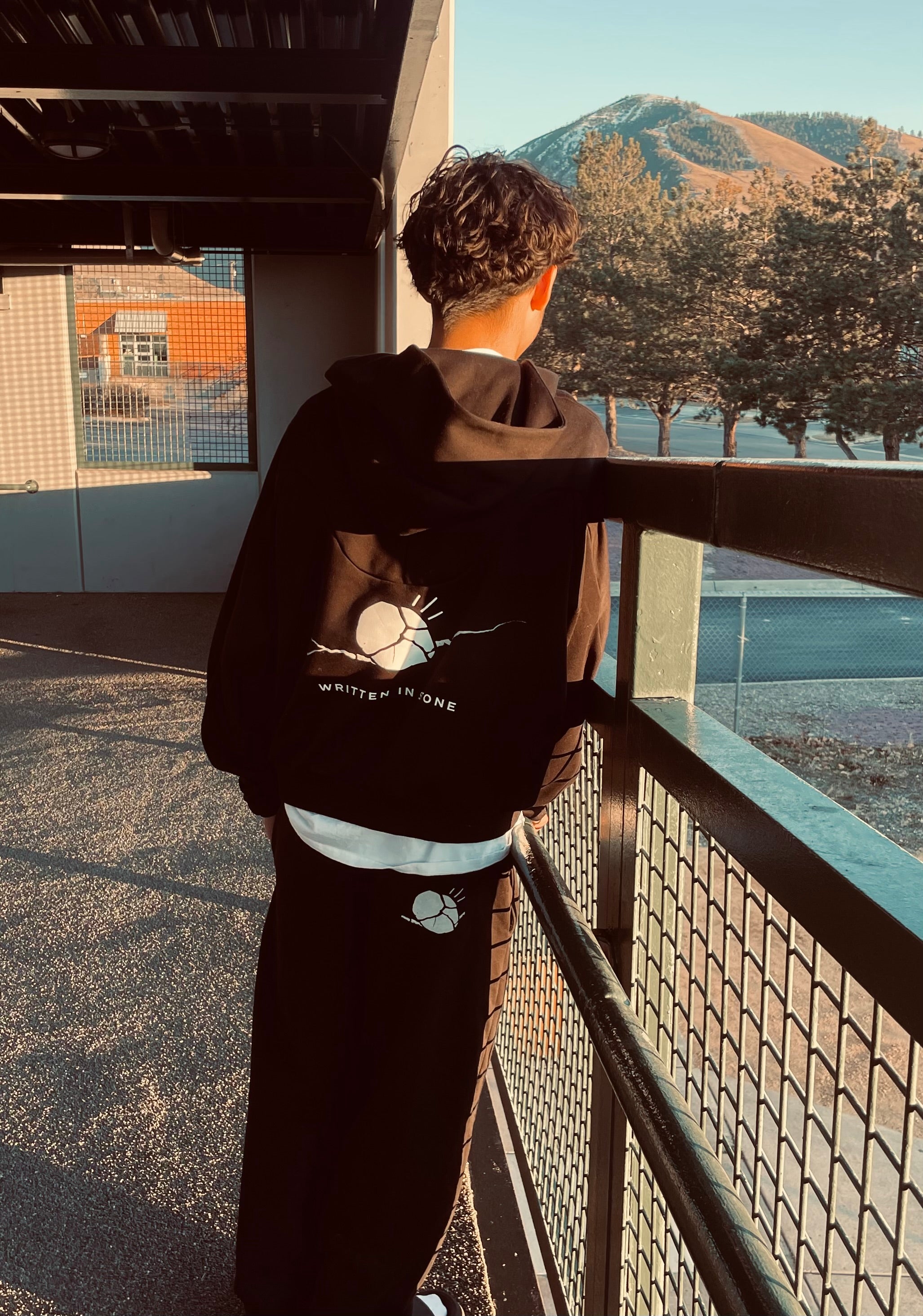 Unisex oversized black raglan hoodie with white "Written in Stone" graphic, worn by a person looking at mountains.