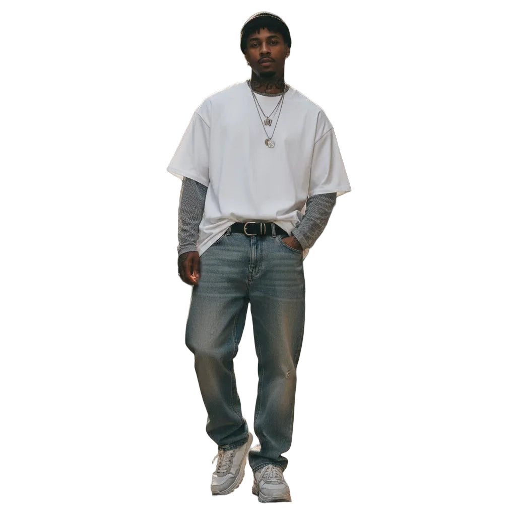 Unisex model wearing a loose white 100% cotton Essential T-Shirt layered over a grey long-sleeve shirt, with light wash jeans and a black beanie, walking in an urban alley.