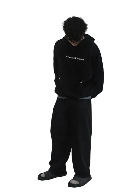 Unisex model wearing a black "STONE CODE" hoodie and loose-fitting black plaid contrast waist pants with gray slides.