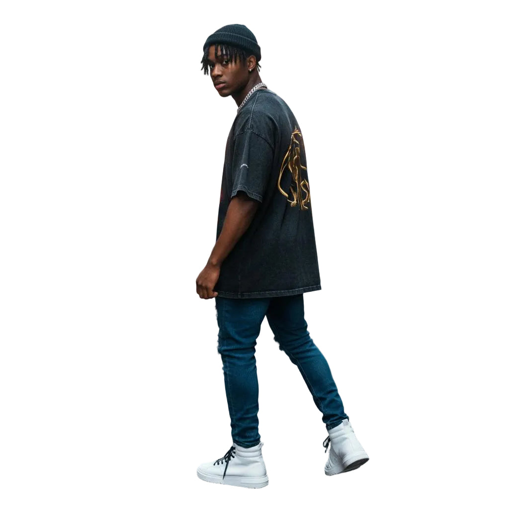 Unisex model wearing a black snow washed oversized cotton t-shirt with a gold graphic, blue jeans, and white sneakers in an alley.